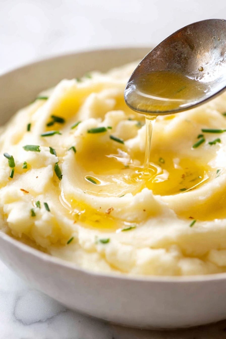 Creamy Mashed Potatoes, mashed potatoes, easy comfort side dish, buttery mashed potatoes, fluffy mashed potatoes - The image shows a close-up of a bowl filled with creamy mashed potatoes that have a smooth, soft texture and a light yellow color. The mashed potatoes are shaped in soft swirls with a slight peak in the center. In the middle, there is a small pool of melted butter that shines with a golden yellow color. Small pieces of green chives are scattered on top, adding contrast and freshness. A shiny silver spoon is dipping into the mashed potatoes on the right side of the bowl. The bowl is white, and it rests on a white marbled surface. photo taken with an iphone --ar 2:3 --v 7