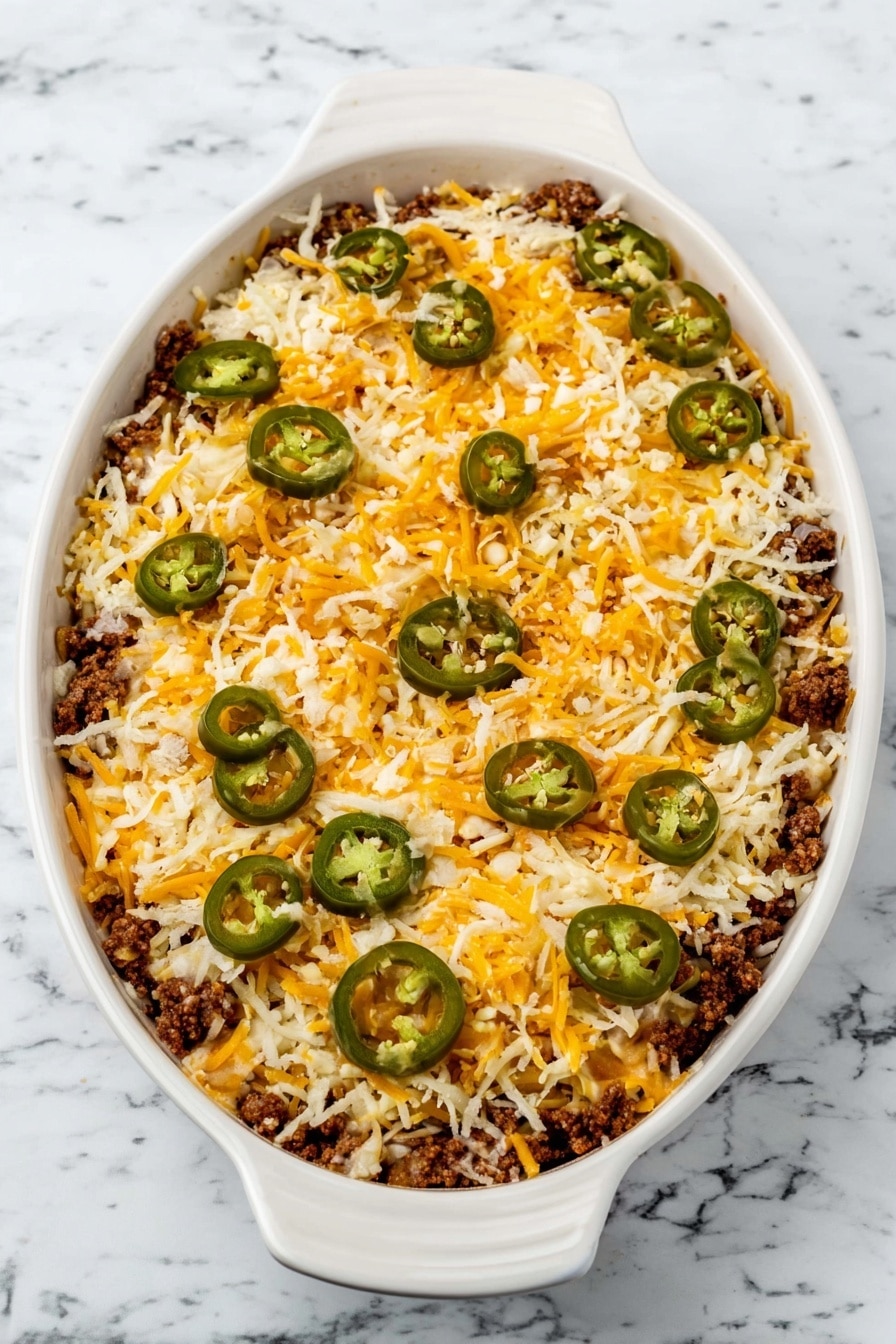 Ground Beef Nachos, cheesy beef nachos, spicy beef nachos, easy nachos recipe, party snack recipes - An oval white ceramic dish with handles is filled with three layers visible from the top view: the bottom layer is a dark brown cooked ground meat, the middle layer is a mix of white and yellow shredded cheese spread evenly, and the top layer is decorated with green sliced jalapeños scattered across the cheese. The dish sits on a white marbled surface. Photo taken with an iphone --ar 2:3 --v 7