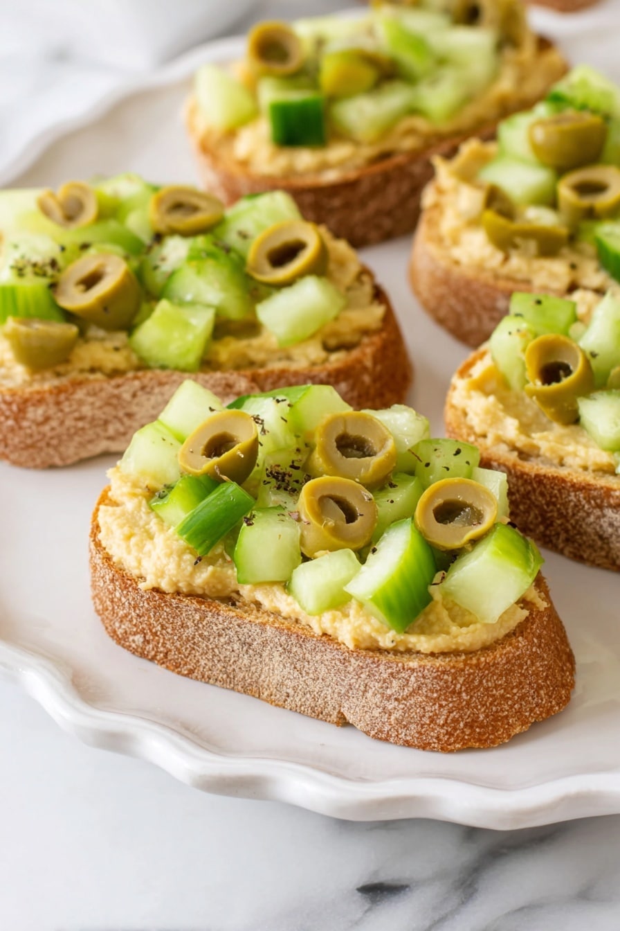 Greek Crostini, Greek Crostini Recipe, Mediterranean Appetizer, Easy Greek Bites, Healthy Crostini Ideas - Small slices of toasted bread form the base layer, with a rough, golden-brown crust and slightly soft inside. On top of each slice, there is a thick spread of creamy, pale yellow hummus, smooth but with small texture spots. The final layer is made up of chopped light green cucumber cubes and sliced green olives, both sprinkled with small black pepper flakes. These small open-faced sandwiches sit on a white plate with scalloped edges, set against a white marbled surface. photo taken with an iphone --ar 2:3 --v 7