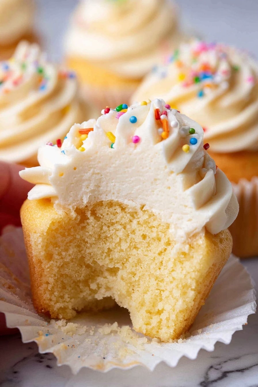 Moist Vanilla Cupcakes, Vanilla Cupcakes Stay Fresh, Fluffy Vanilla Cupcakes, Easy Vanilla Cupcake Recipe, Long-Lasting Vanilla Cupcakes - A close-up of a vanilla cupcake with one layer of golden cake at the bottom, wrapped in a white paper liner. On top, there is a thick swirl of smooth, creamy off-white frosting being piped from a metal tip with a clear piping bag, creating peaked curves. Around the main cupcake, three more cupcakes are visible, two with similar frosting swirls and one without frosting, all resting on a white marbled surface. photo taken with an iphone --ar 2:3 --v 7