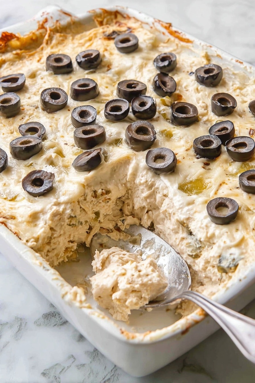Knock-Your-Socks-Off Warm Crab Dip, crab dip appetizer, creamy crab dip recipe, crowd-pleasing seafood dip, easy warm crab dip - This is a white baking dish filled with a creamy, thick casserole that has a smooth, slightly browned top layer. On top, there is a scattered layer of black olive slices, each slice dark with a shiny texture. Underneath the top, thick layers of creamy mixture with chunks are visible where a portion has been scooped out, showing a soft, mixed texture in pale beige and light greenish tones. A silver spoon rests in the dish, partially covered with the casserole. The dish is placed on a white marbled surface. photo taken with an iphone --ar 2:3 --v 7