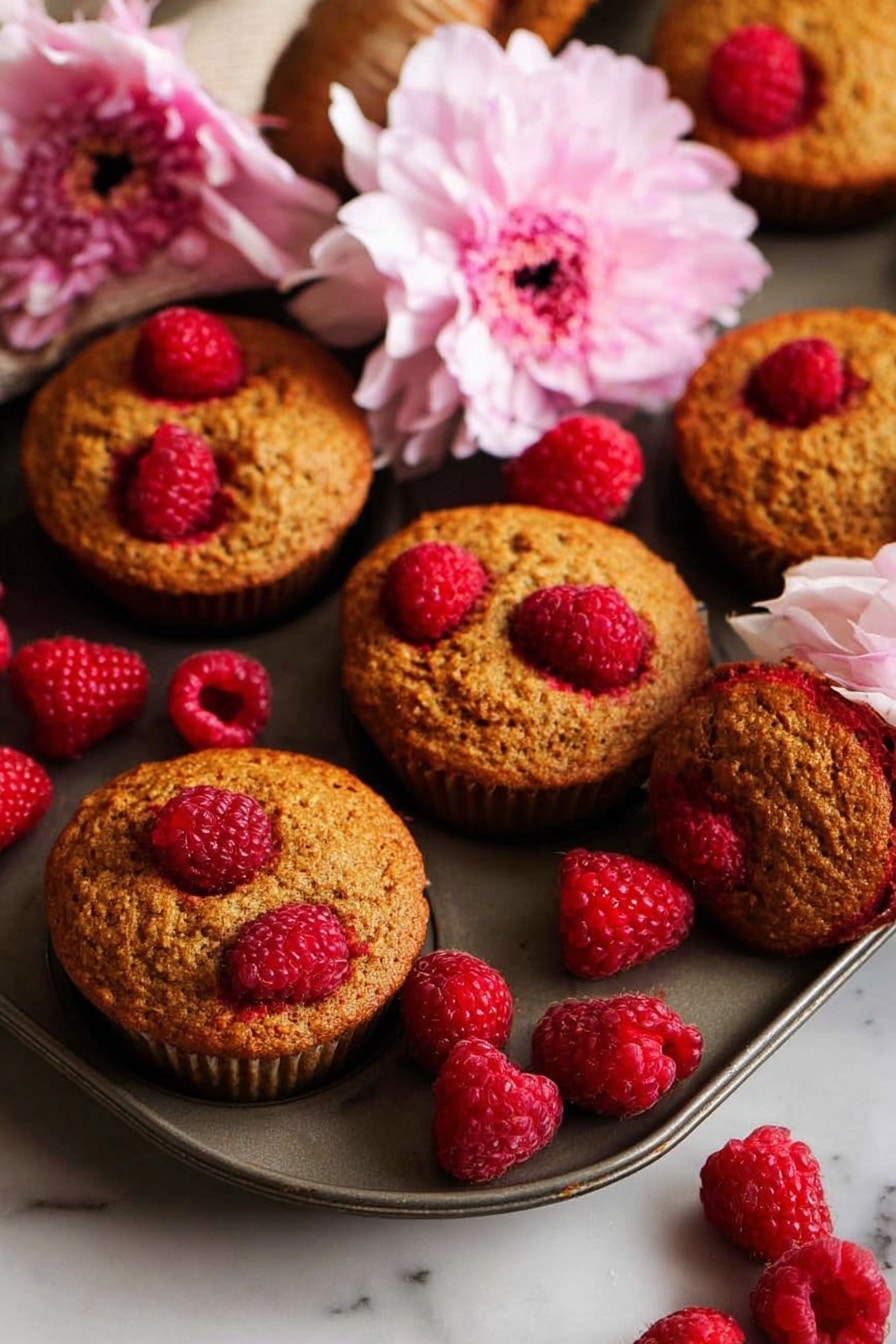 Up-and-Go Breakfast Muffins with Raspberries and Whole Wheat Flour, healthy raspberry breakfast muffins, quick wholesome breakfast recipes, nutritious muffin ideas, raspberry whole wheat muffins - The image shows a tray with eight golden-brown muffins, each topped with bright red raspberries that create a contrast on the warm textured muffin tops. Several extra fresh raspberries are scattered around the tray, adding vibrant red color and moist texture. A large pink flower rests near the muffins, offering a soft and delicate touch against the rough muffin surface. The scene sits on a white marbled surface that gives a clean and bright backdrop. The whole setup is warmly lit, emphasizing the rich colors and textures of the muffins and raspberries. photo taken with an iphone --ar 2:3 --v 7