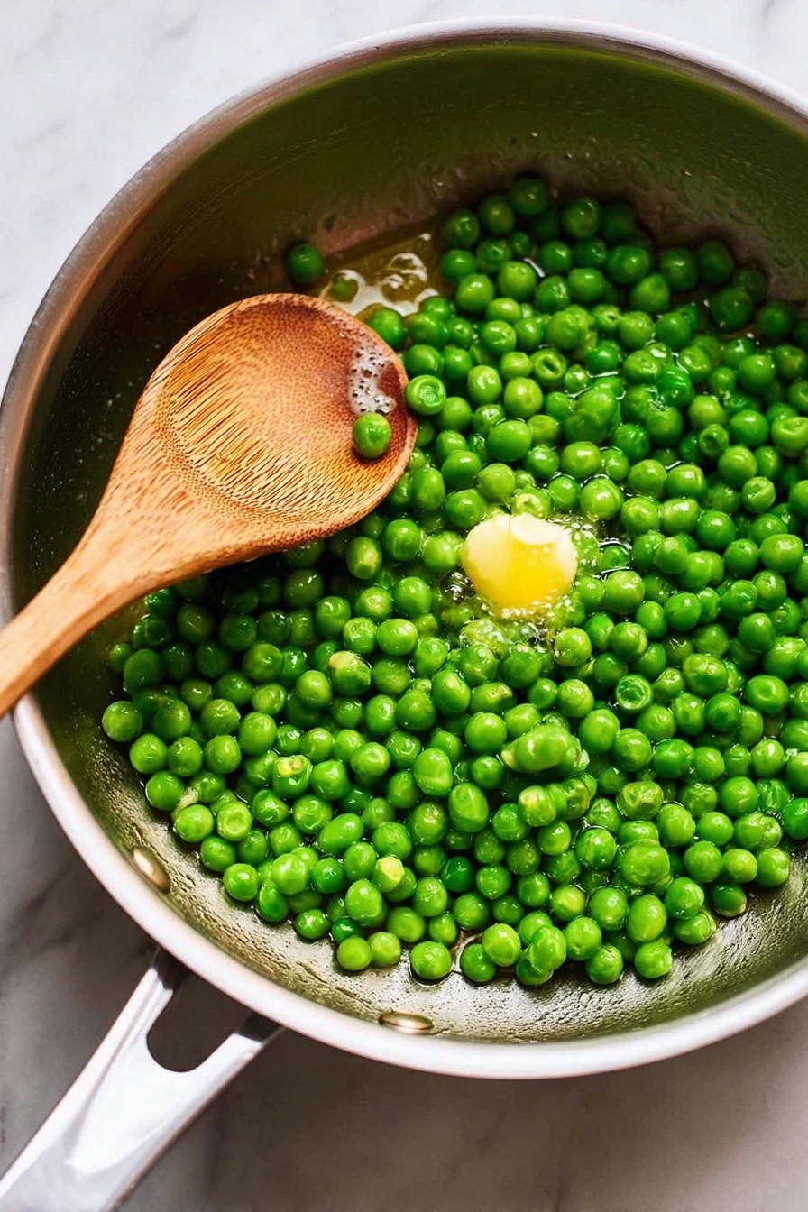 Garlic Buttered Peas, garlic butter peas, quick green vegetable side, easy peas side dish, healthy vegetable recipes - The image shows a close-up of a white bowl filled with bright green peas that look shiny and fresh. The peas are packed closely together, and you can see small bits of what looks like seasoning or herbs scattered on top. A silver spoon is partly dipped into the peas, resting in the bowl at the right side with its handle stretching out. The background is a white marbled texture that adds a clean, soft feel to the image. Photo taken with an iphone --ar 2:3 --v 7