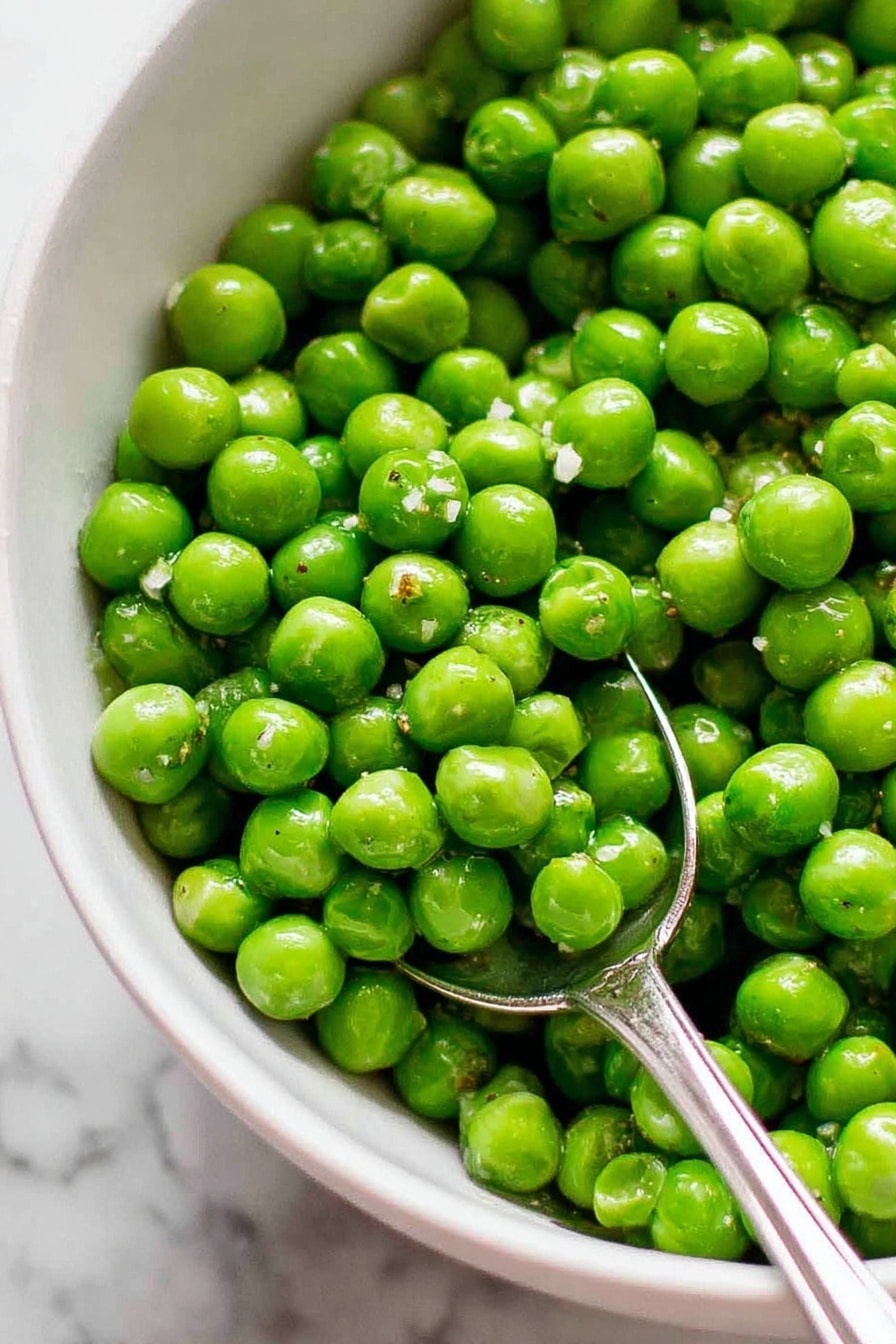 Garlic Buttered Peas, garlic butter peas, quick green vegetable side, easy peas side dish, healthy vegetable recipes - A large silver pan filled with bright green peas as the main layer, with a small dollop of yellow butter melting on top. A wooden spoon is stirring the peas from the left side, showing a mix of smooth green textures and slight shiny spots from the heat. The pan sits on a white marbled surface, adding a clean and simple background. Photo taken with an iphone --ar 2:3 --v 7