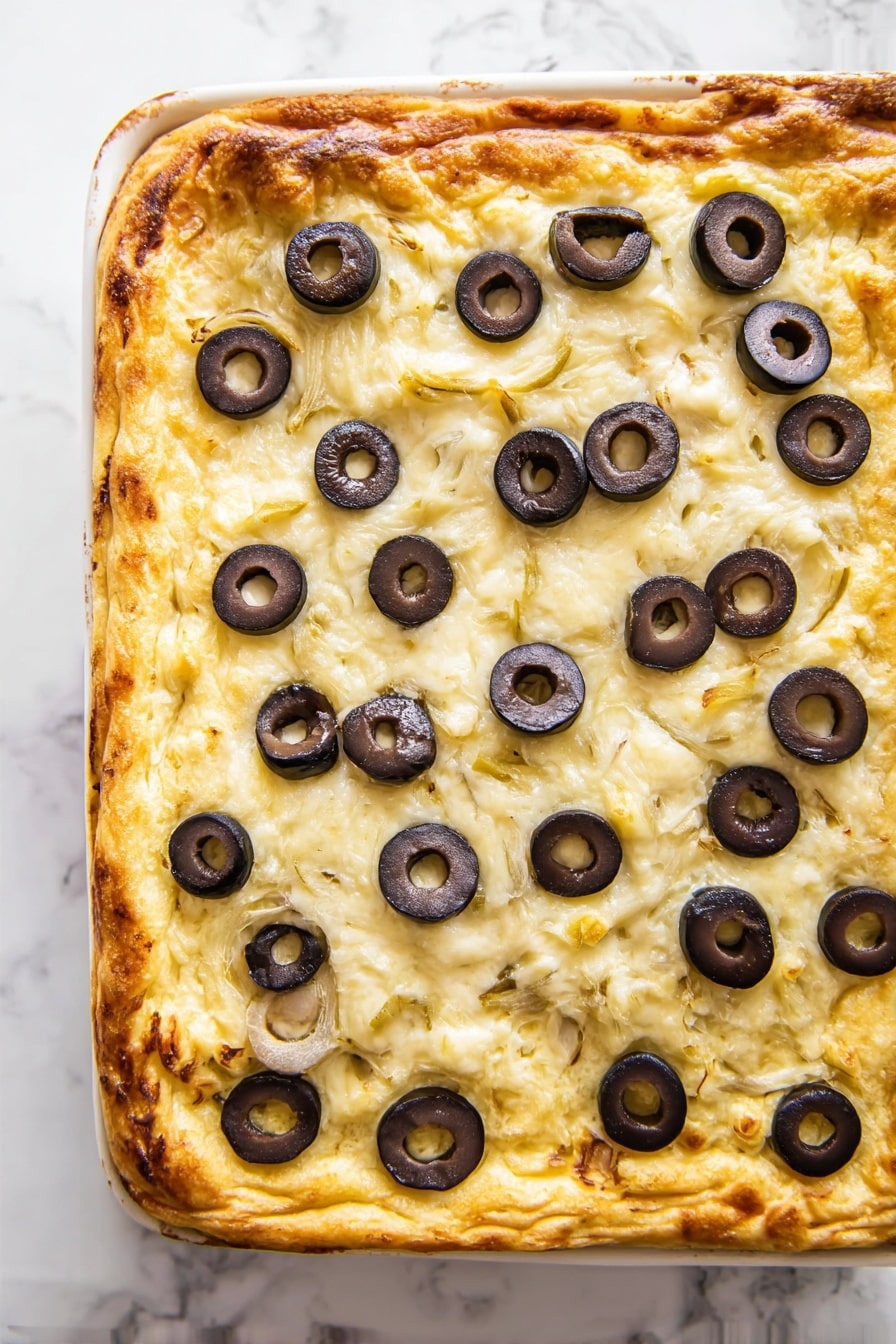 Knock-Your-Socks-Off Warm Crab Dip, crab dip appetizer, creamy crab dip recipe, crowd-pleasing seafood dip, easy warm crab dip - A square flat dish with one thick golden-brown baked layer forming the crust, topped with a creamy light beige cheese layer that covers the entire surface. Scattered evenly on top are many sliced black olives with dark outer edges and a lighter inner ring, adding contrast. The edges of the crust show a slightly darker browned color, indicating a crisp texture. The dish is on a white marbled surface. photo taken with an iphone --ar 2:3 --v 7