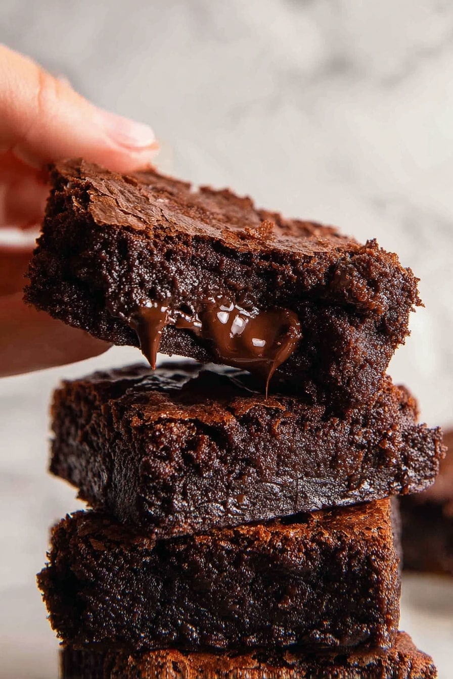 Easy Chocolate Brownies, fudgy brownies, chocolate brownie recipe, quick brownie ideas, homemade chocolate desserts - A close-up view of three stacked chocolate brownies with a rich, dark brown color. The top layer has a slightly cracked, dry crust, while the inside looks moist and melted with gooey chocolate chunks visible in the middle and towards the edges. The brownies have a soft texture with irregular edges, and the bottom one sits on a white marbled surface with crumbs scattered around. photo taken with an iphone --ar 2:3 --v 7