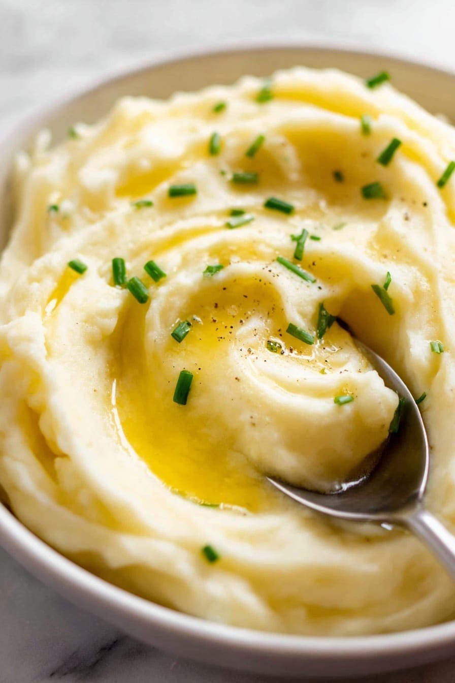 Creamy Mashed Potatoes, mashed potatoes, easy comfort side dish, buttery mashed potatoes, fluffy mashed potatoes - A close-up view shows creamy mashed potatoes in a white bowl with a soft, smooth texture and some small lumps for a natural look. The surface has a gentle swirl pattern with small green chive pieces sprinkled on top. A shiny silver spoon held by a woman's hand is pouring golden melted butter in a thin stream, creating a glossy wet layer on part of the potatoes. The background has a white marbled texture with soft lighting that highlights the butter shine and creamy potato surface. photo taken with an iphone --ar 2:3 --v 7