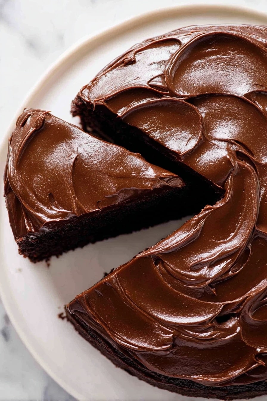 Easy Chocolate Fudge Cake, chocolate fudge cake, rich chocolate dessert, simple chocolate cake, indulgent chocolate treat - A close-up view of a round, single-layer chocolate cake covered entirely with thick, smooth, and shiny dark chocolate frosting that has curved swirls and waves. A slice is slightly separated from the whole cake, showing the rich dark color of the cake underneath. The cake sits on a white plate, placed on a white marbled surface. Photo taken with an iphone --ar 2:3 --v 7