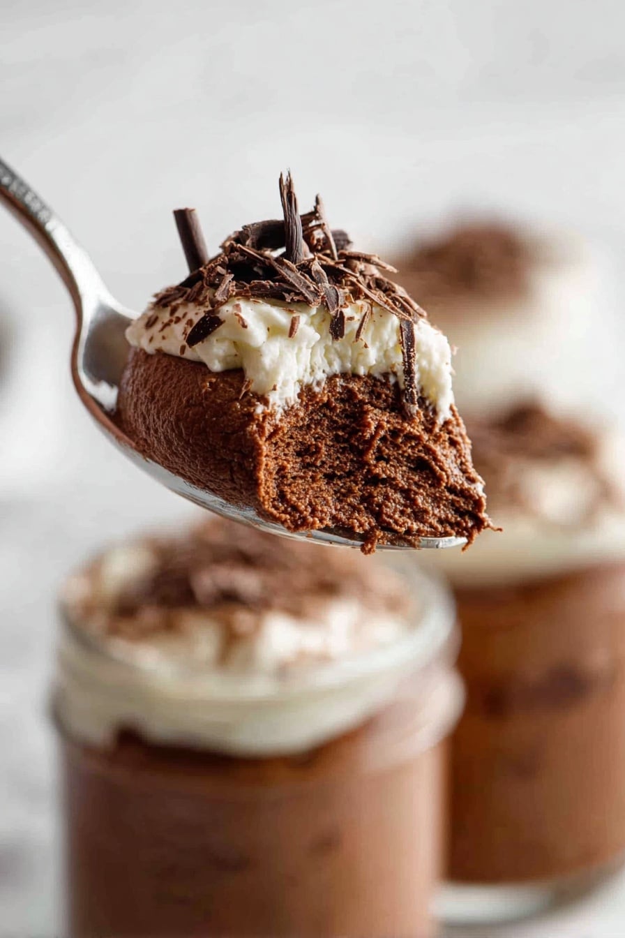 Decadent Chocolate Mousse, Chocolate Mousse Recipe, Easy Chocolate Dessert, Silky Chocolate Dessert, Rich Chocolate Treats - A close-up of a spoon holding a fluffy, dark brown chocolate mousse layer with a light, creamy white topping adorned with thin dark chocolate shavings. The spoon is lifted above two white jars filled with the same layered dessert, showing the mousse's airy texture and smooth whipped cream on top. The background has a soft, white marbled texture that highlights the rich chocolate colors. Photo taken with an iphone --ar 2:3 --v 7