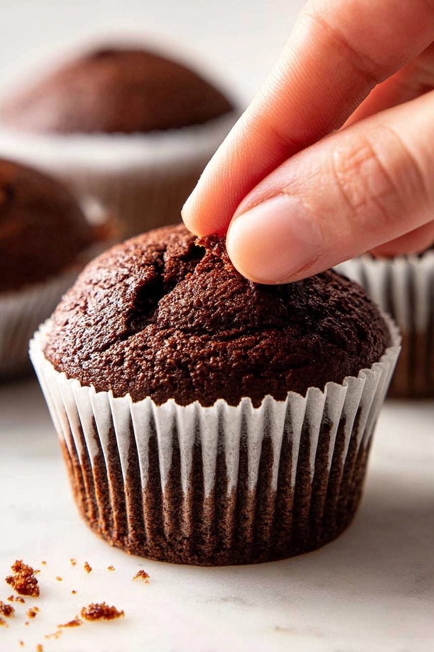 Vanilla Cream Filled Chocolate Ganache Cupcakes, chocolate cupcakes with vanilla cream filling, decadent chocolate cupcakes with ganache, homemade vanilla cream cupcakes, rich chocolate dessert cupcakes - A close-up view of a dark brown chocolate muffin with a cracked top, sitting inside a white paper liner with ridges around the edge. A woman's finger is gently pressing the soft surface of the muffin, showing its texture. The muffin rests on a white marbled surface with small crumbs scattered around. In the blurred background, two more chocolate muffins in white paper liners can be seen. Photo taken with an iphone --ar 2:3 --v 7