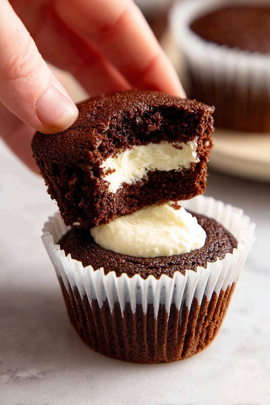 Vanilla Cream Filled Chocolate Ganache Cupcakes, chocolate cupcakes with vanilla cream filling, decadent chocolate cupcakes with ganache, homemade vanilla cream cupcakes, rich chocolate dessert cupcakes - A close-up image shows a chocolate cupcake with a white paper liner sitting on a white marbled surface. The cupcake has three visible layers: the bottom dark brown cake base, a smooth white cream filling in the middle, and a top dark brown cake piece being lifted by a woman's hand. The cream filling looks soft and thick, sitting visibly inside the cupcake cavity. In the background, there is another similar cupcake slightly out of focus. Photo taken with an iphone --ar 2:3 --v 7